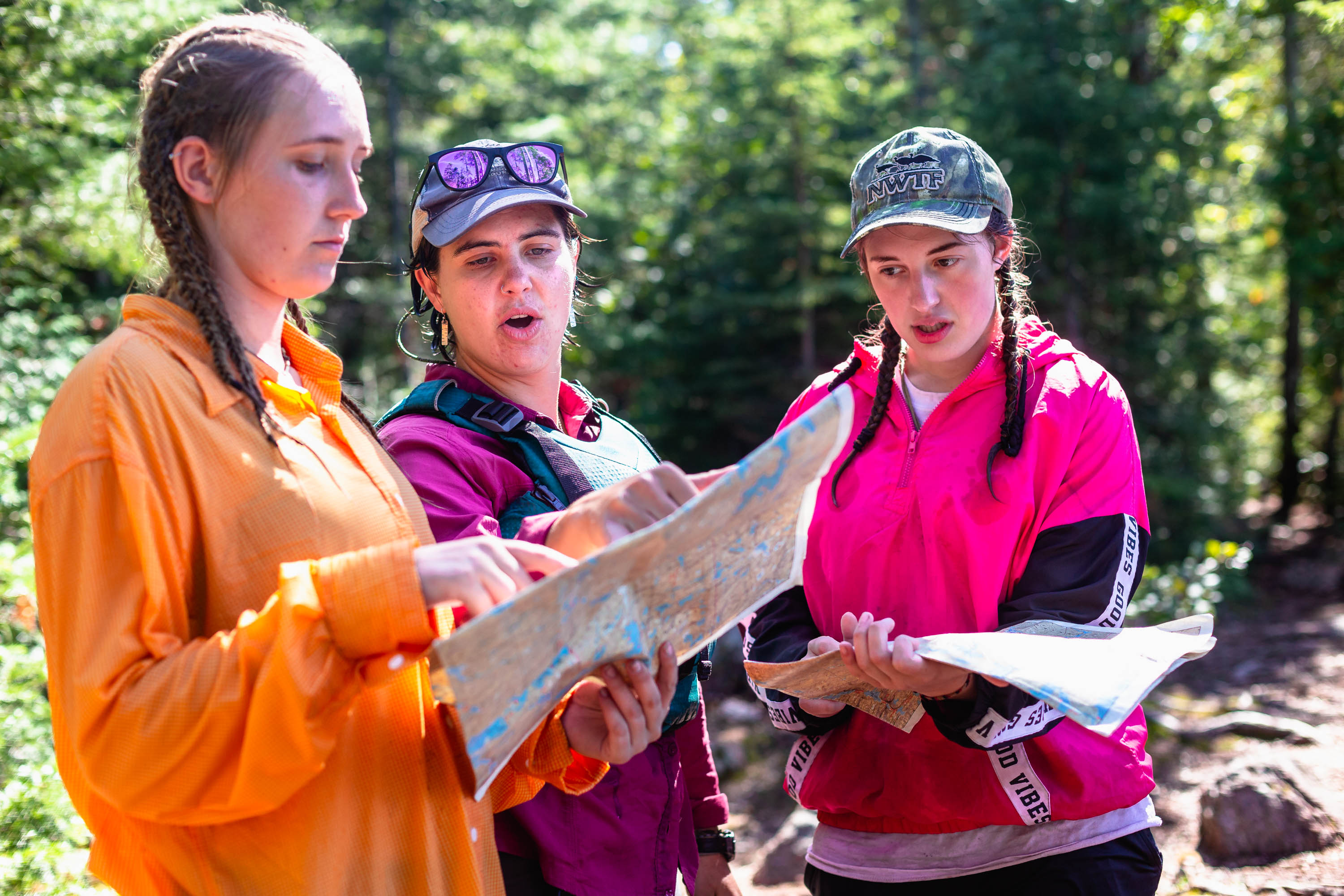 Three young women are looking at a map in a forest. The woman on the left is wearing an orange shirt and pointing at the map. The woman in the middle is wearing a purple shirt and a baseball cap. The woman on the right is wearing a pink jacket and a baseball cap. They appear to be hiking or camping.